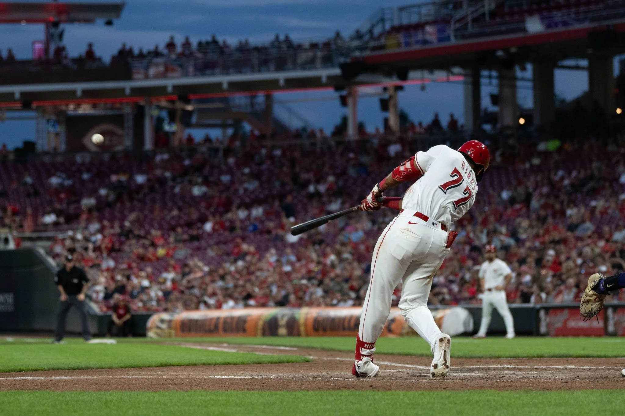 A baseball player in a white uniform and red helmet is swinging a bat at a pitch during a game, with a packed stadium in the background.