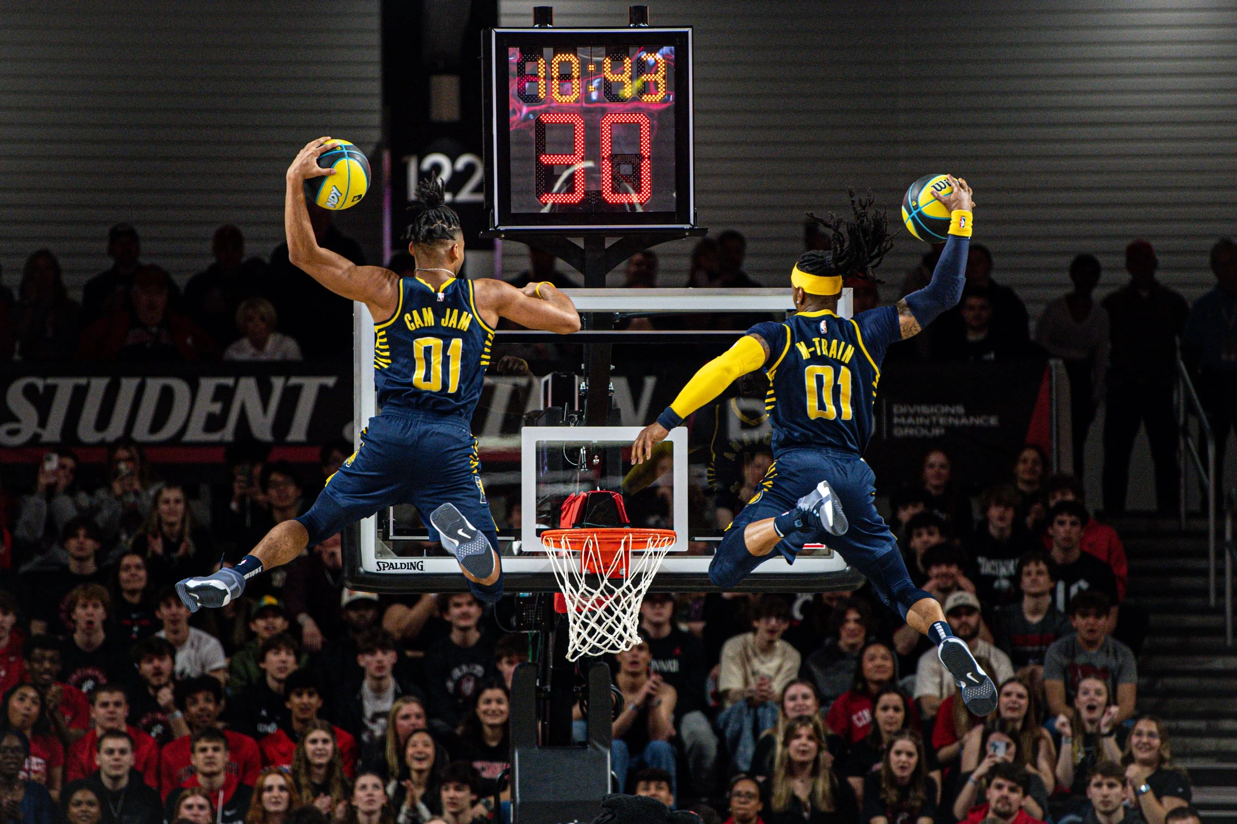 Two basketball players wearing matching jerseys with the number 01 and the names 'Cam Jam' and 'M-Train' are performing a dunk during a game, with an audience watching in the background.