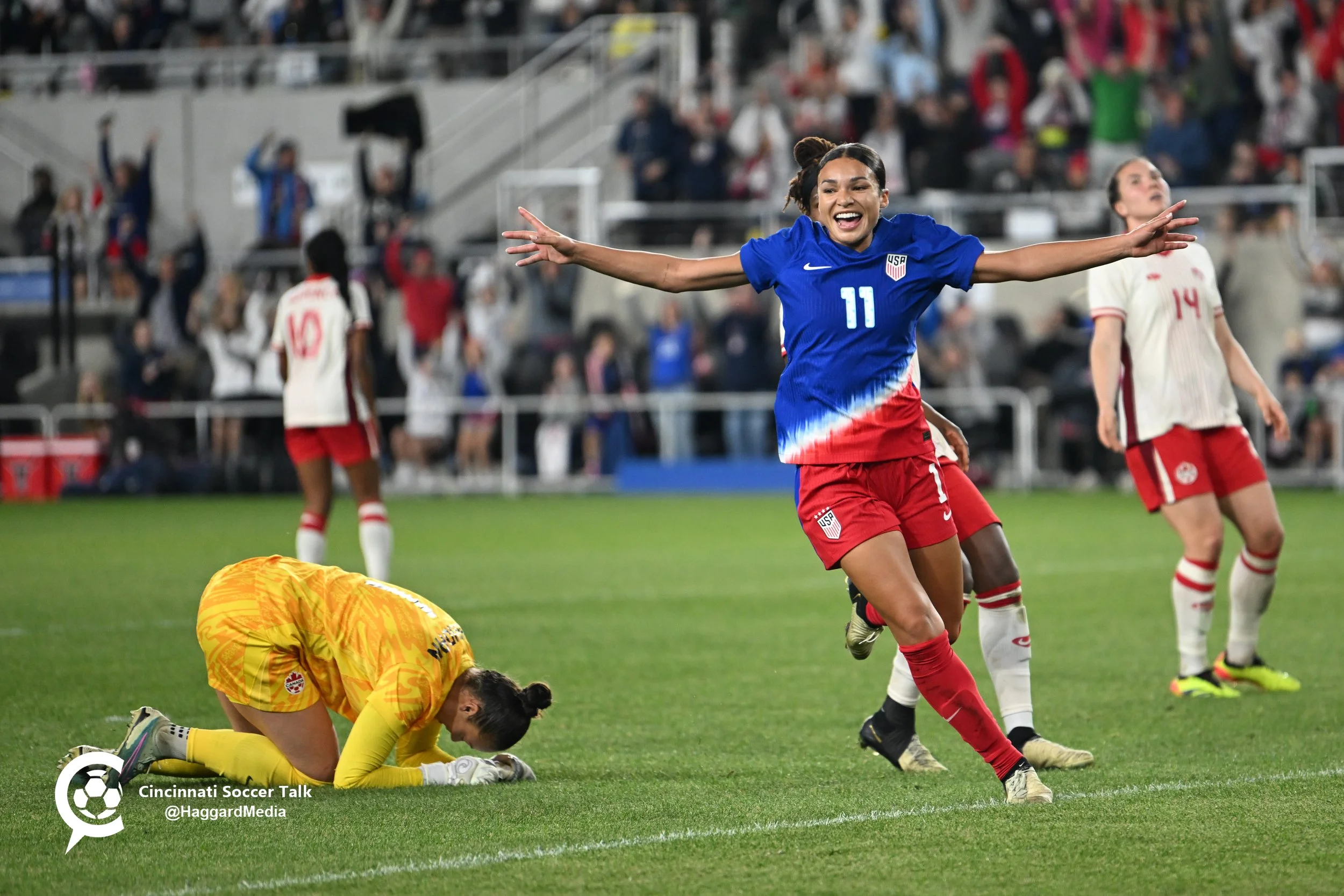 A female soccer player in a blue and red uniform celebrating on the field with arms outstretched, while other players and a goalkeeper kneel or stand nearby during a match in a stadium filled with spectators.