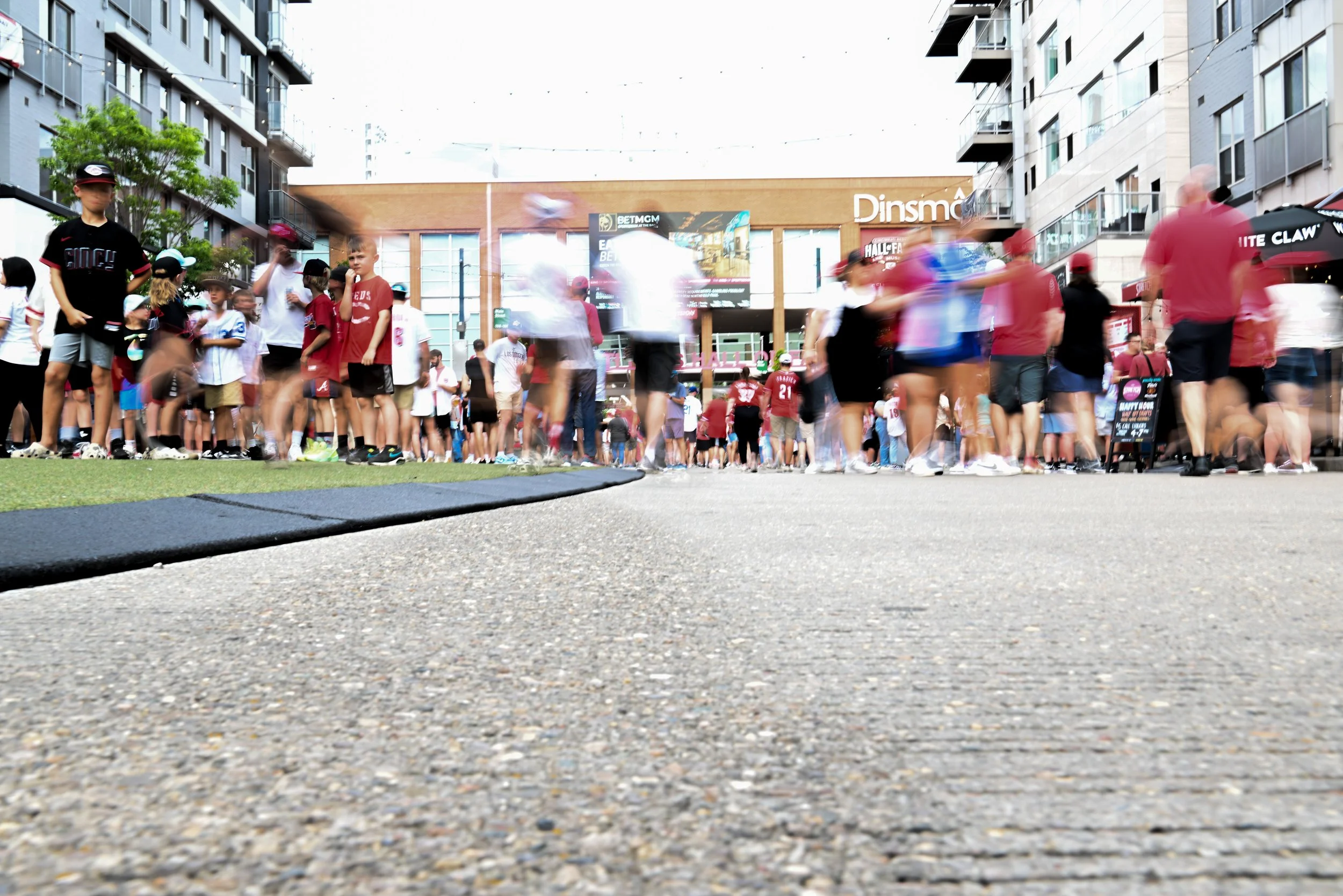 Crowd of people standing in line outdoors on a city street, with buildings and storefronts in the background, some motion blur indicating movement.