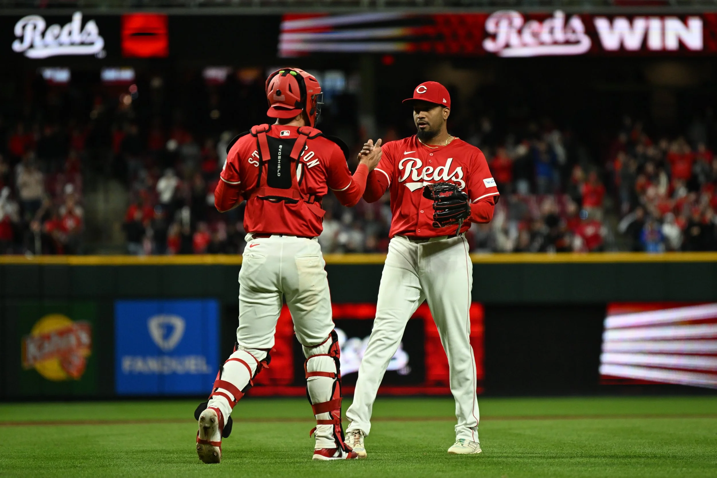 Two baseball players in red uniforms with 'Reds' written on the front, standing on the field and exchanging a fist bump during a game, with crowd and stadium in the background.