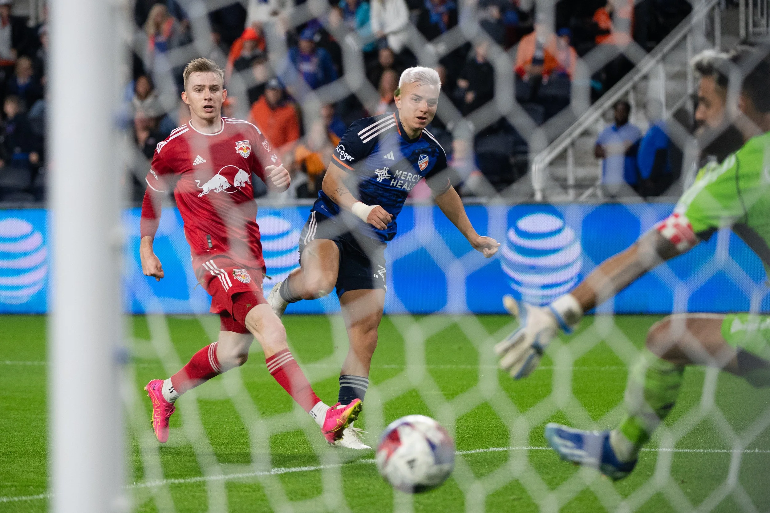 A soccer match FC Cincinnati and New York Red Bulls with two players chasing the ball near the goal, with a goalkeeper reaching out to block it. Spectators are visible in the background.