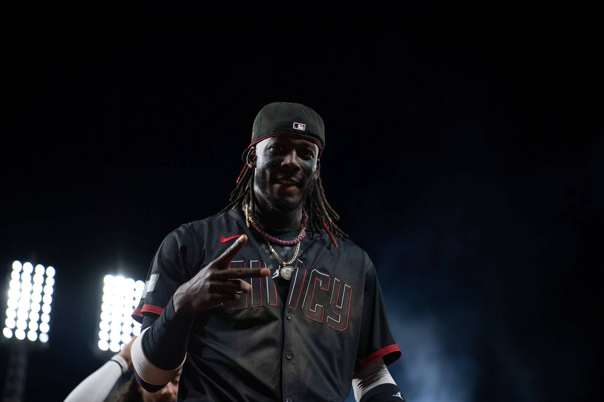 A man wearing a black baseball uniform with red accents, a black hat with a sports logo, and jewelry, making a peace sign with his right hand, standing under stadium lights at night.