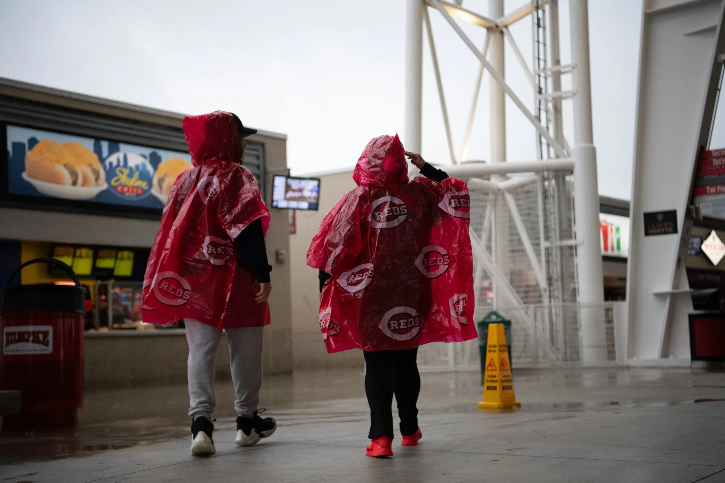 Three people in red Cincinnati Reds rain ponchos walking inside a stadium concourse.