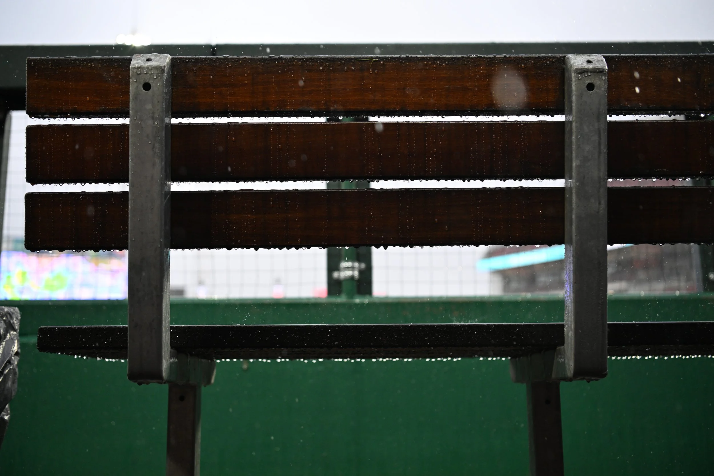 A park bench made of dark wooden slats with metal supports, wet from rain, on a rainy day with some raindrops falling.