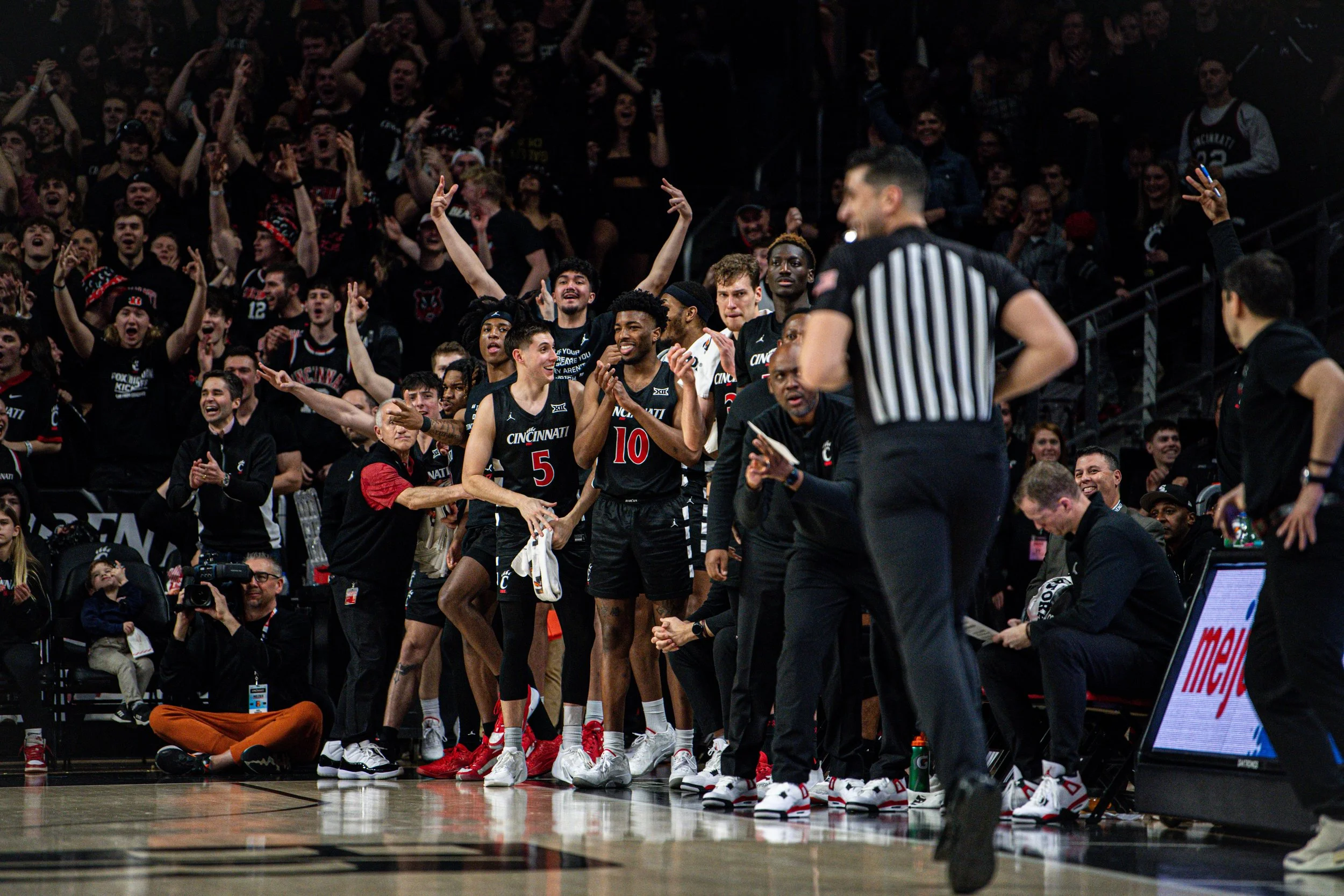 Uptempo basketball game with players and fans celebrating on the sidelines, some wearing black jerseys with red and white accents, in a crowded indoor arena.