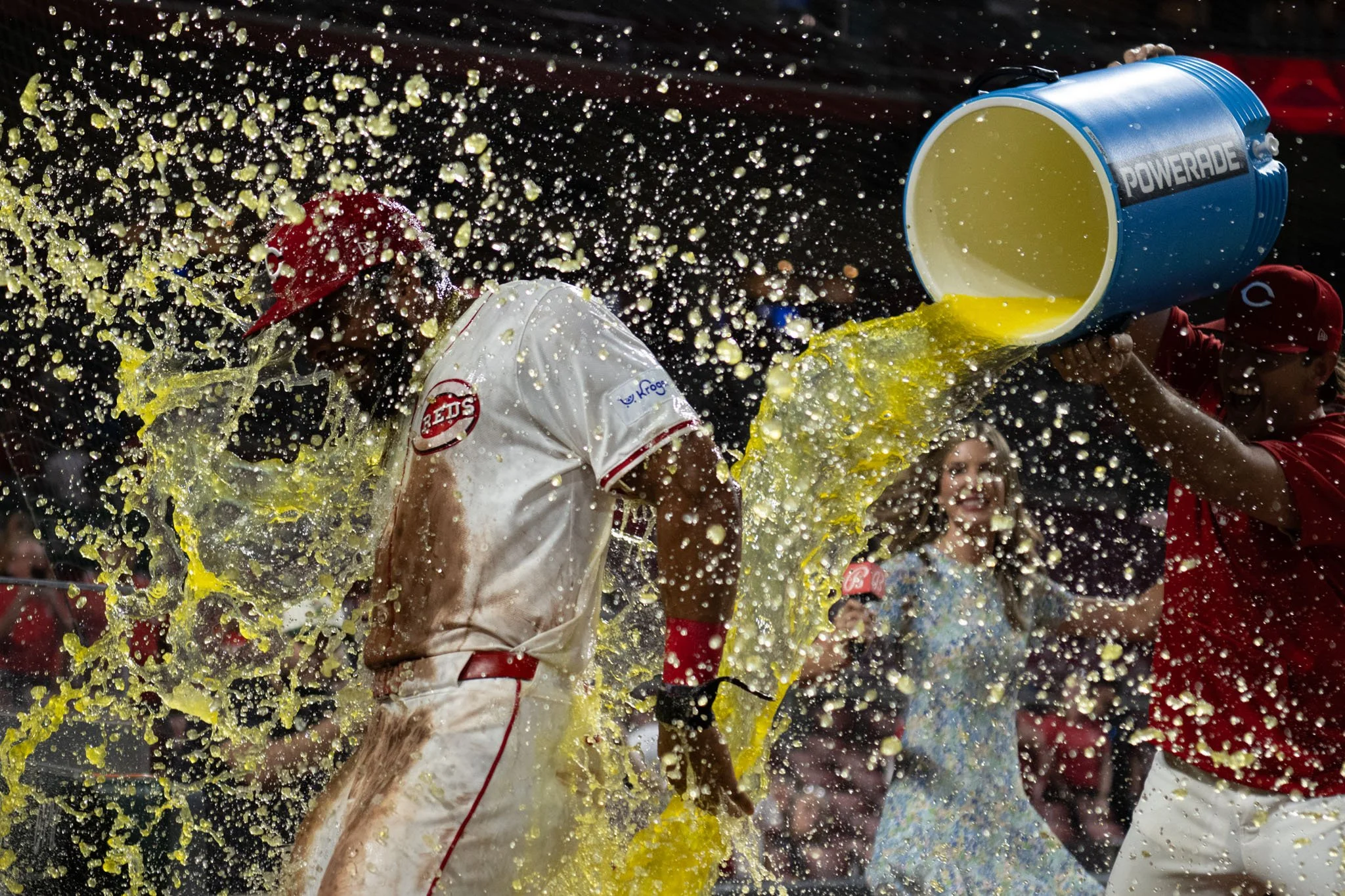 A person in a white baseball uniform and red cap getting drenched with yellow-orange sports drink poured from a large blue Gatorade container by another person in a red shirt, with a woman in the background smiling.