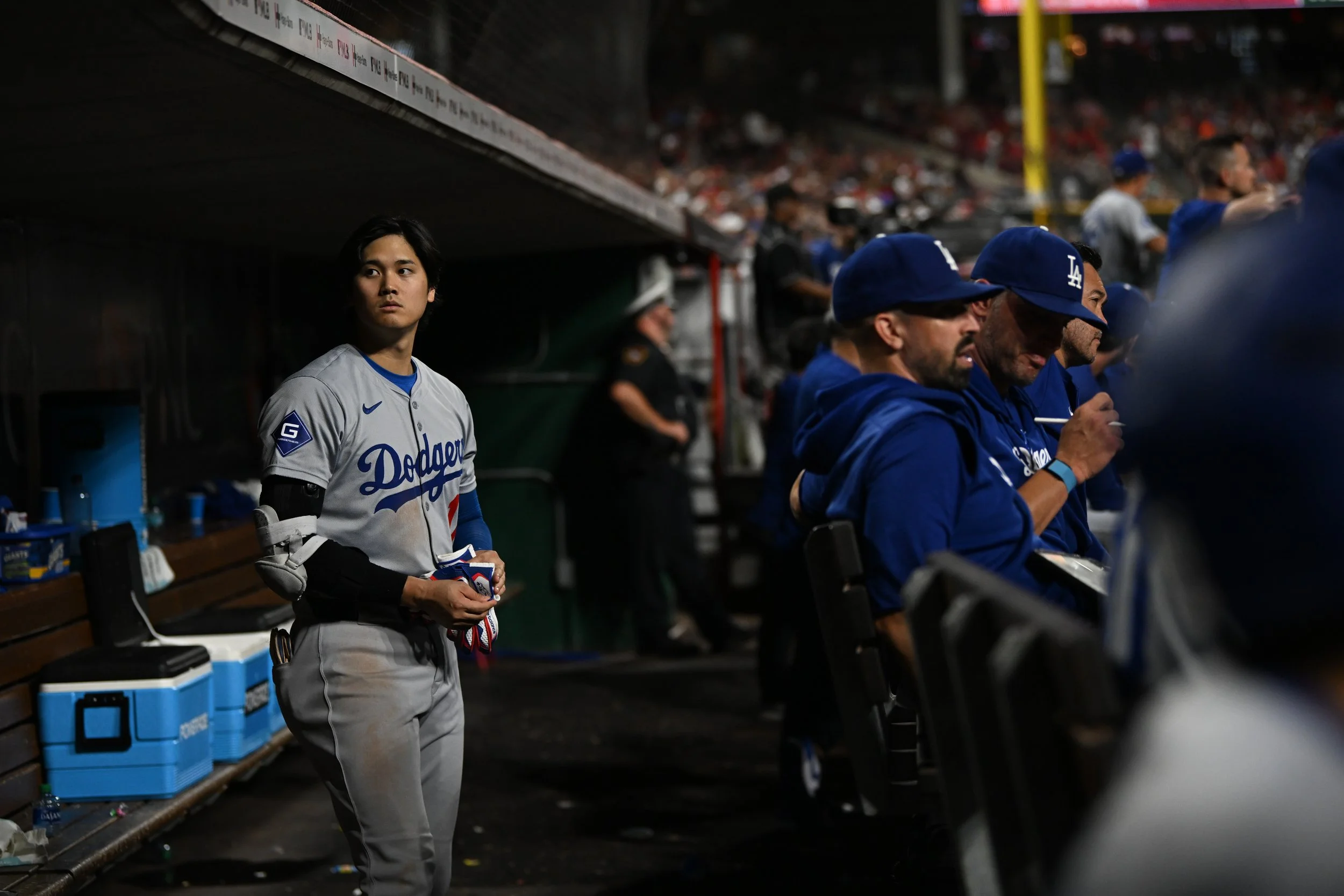 A baseball player in a gray Los Angeles Dodgers uniform standing in the dugout, holding a glove, with other team members seated nearby and a crowd in the background.