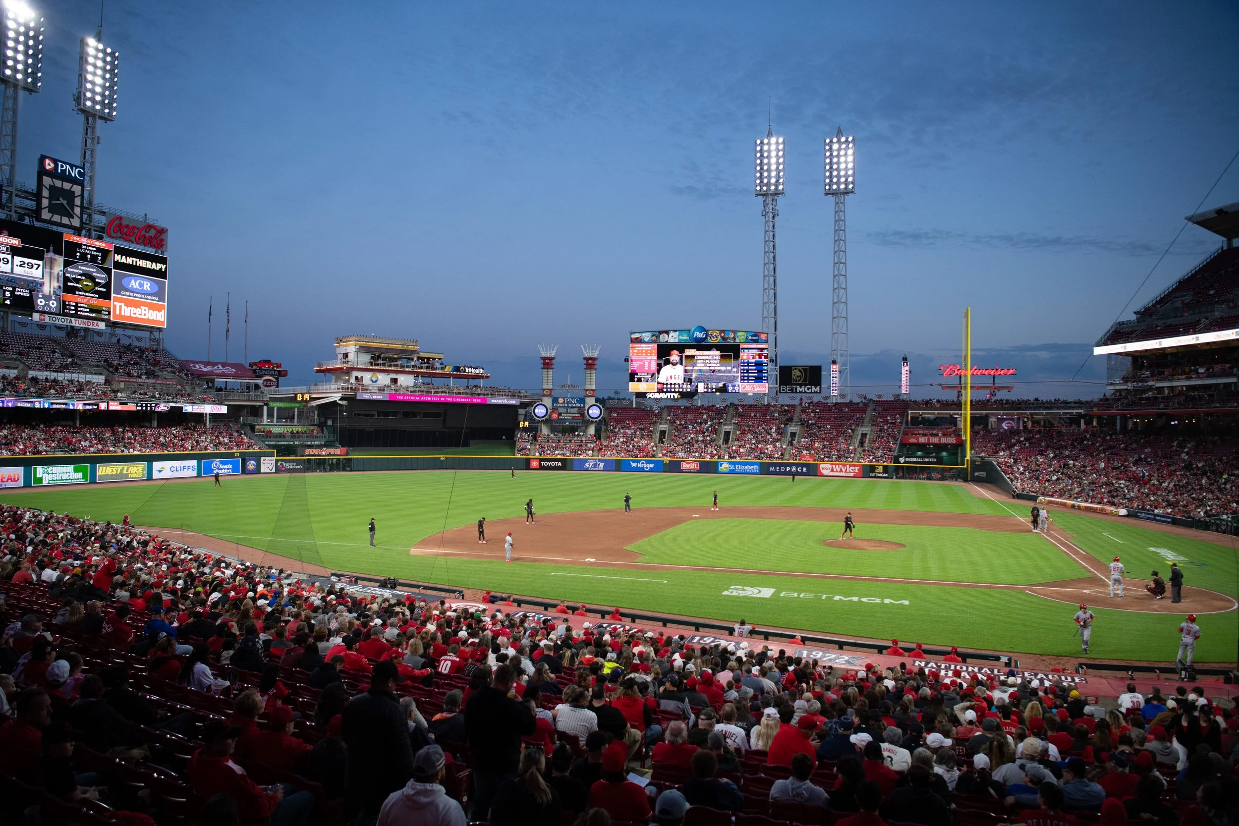A baseball game in progress at a stadium during evening with crowd in stands, players on the field, and large digital scoreboard in the background.