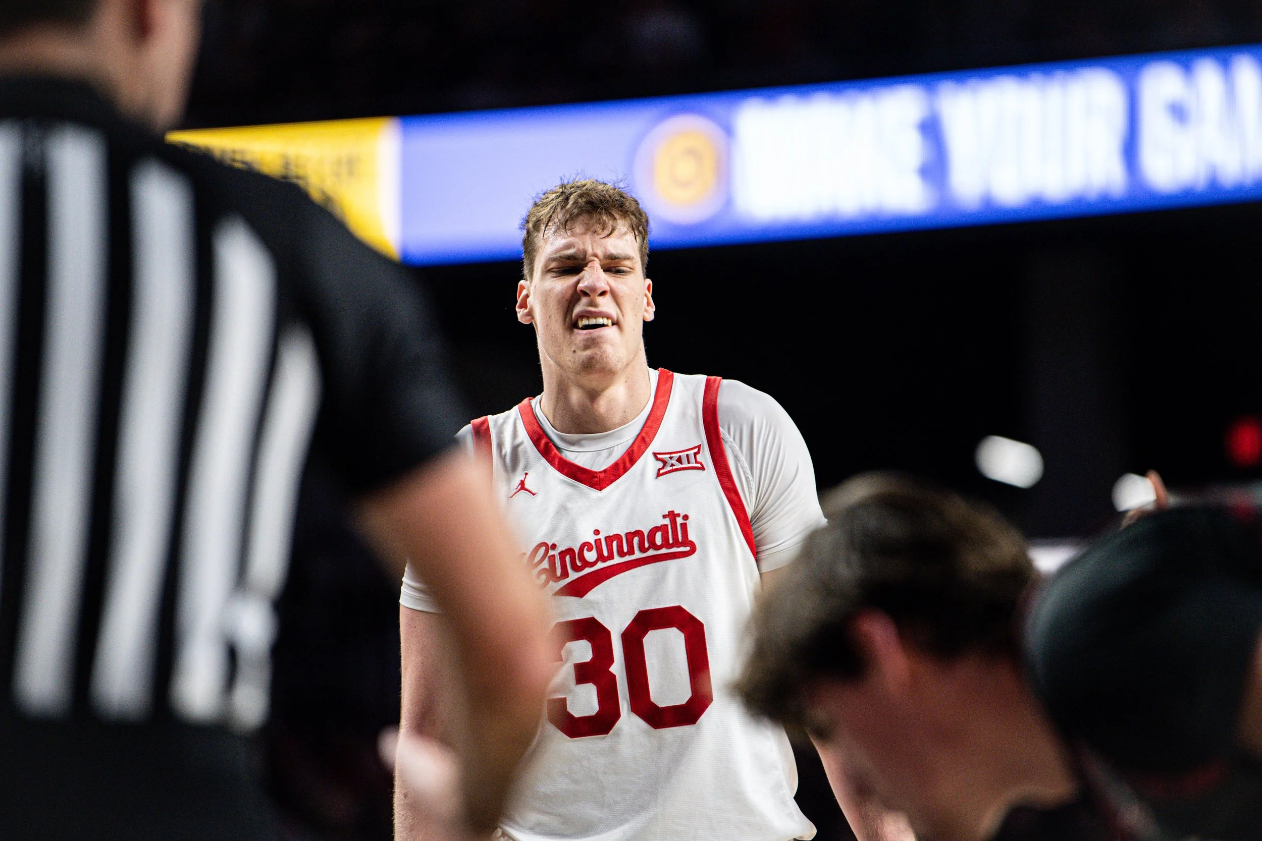 An intense moment during a basketball game featuring a University of Cincinnati player wearing jersey number 30, showing discomfort or pain, with referees and other players visible around.
