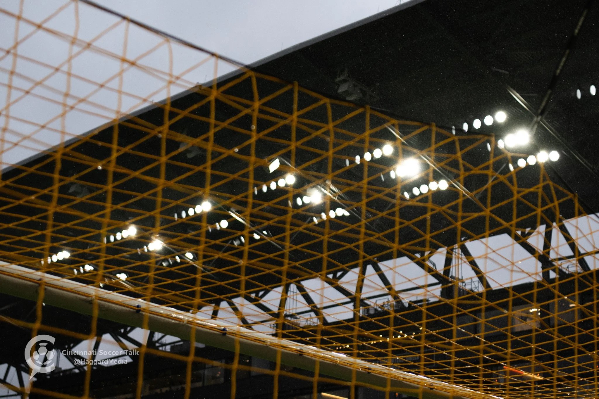 A soccer goal net with a stadium and bright lights in the background.