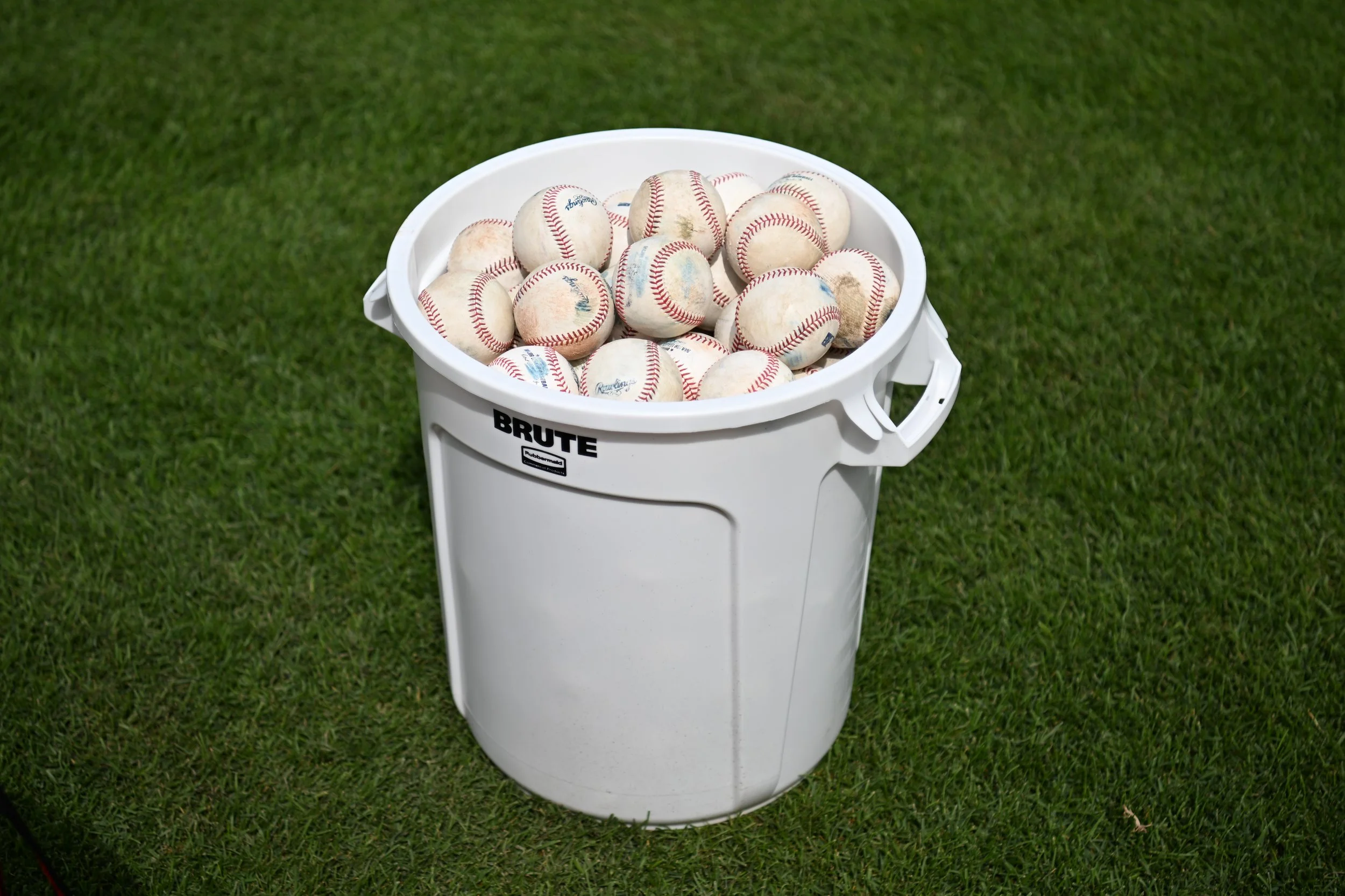 A white bucket filled with used baseballs sitting on a green grass field.