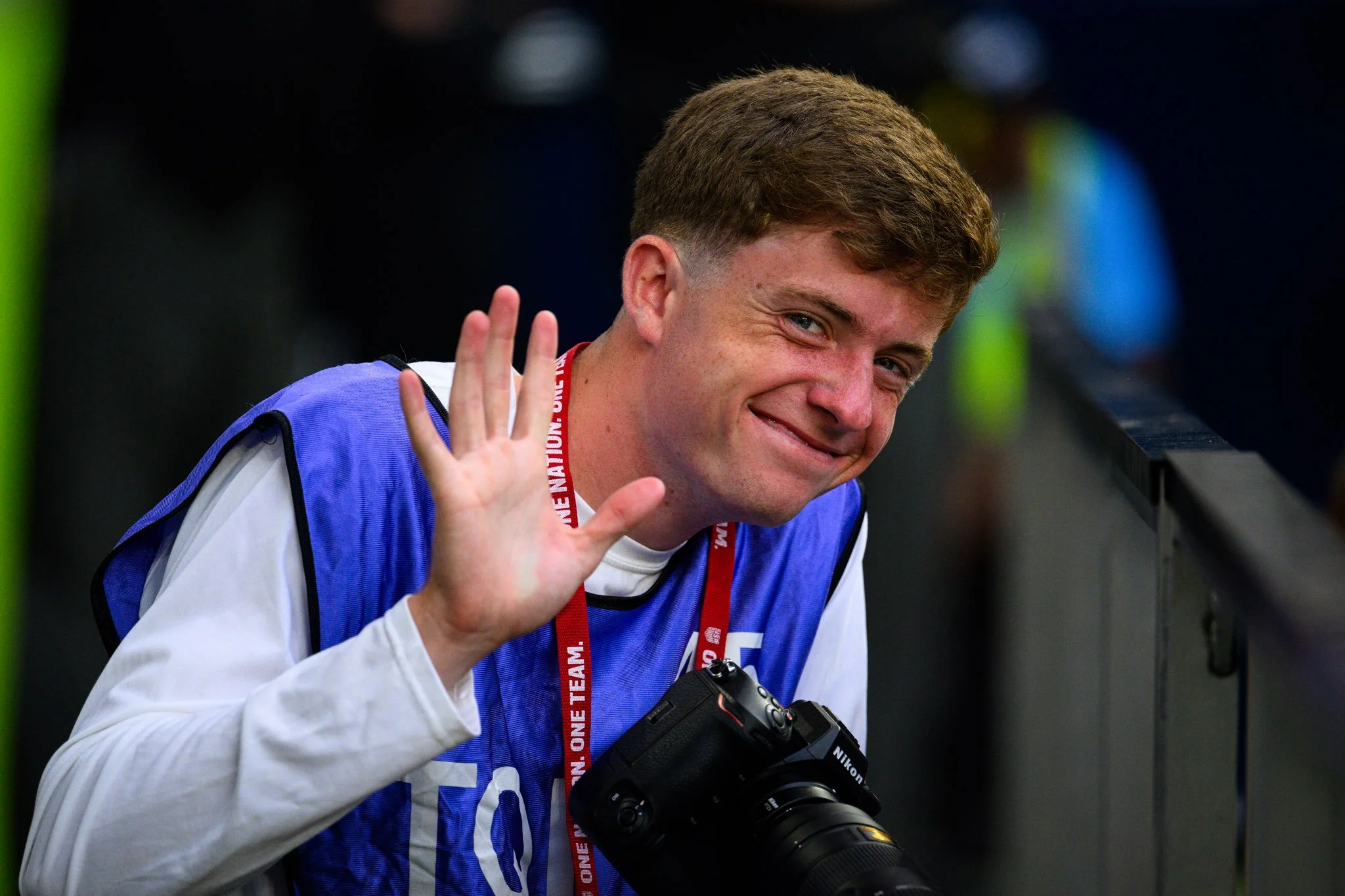 A young man with brown hair smiling and waving, wearing a blue vest over a white shirt, with a Nikon camera hanging around his neck, at an indoor event.