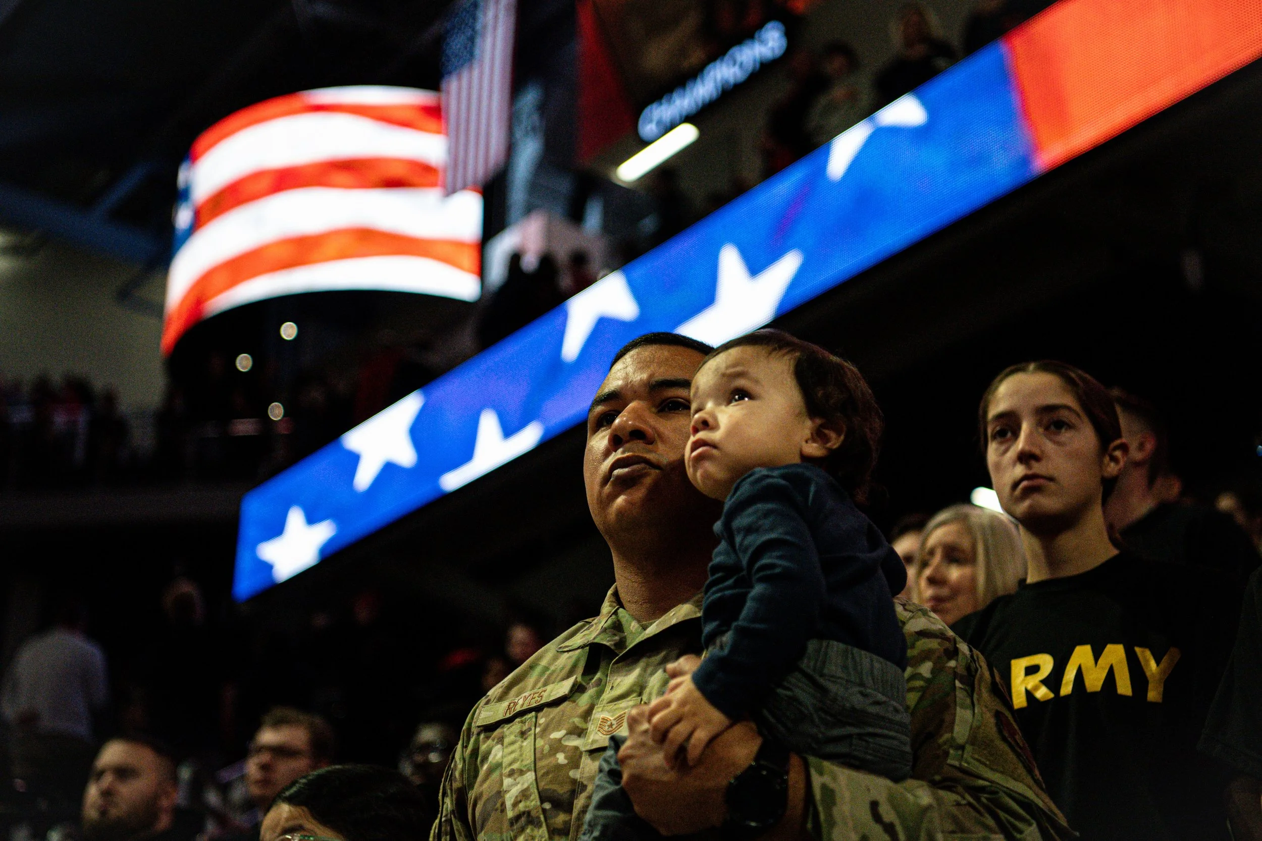 A soldier holding a young child at a patriotic event, with American flags and a large display of stars and stripes in the background.