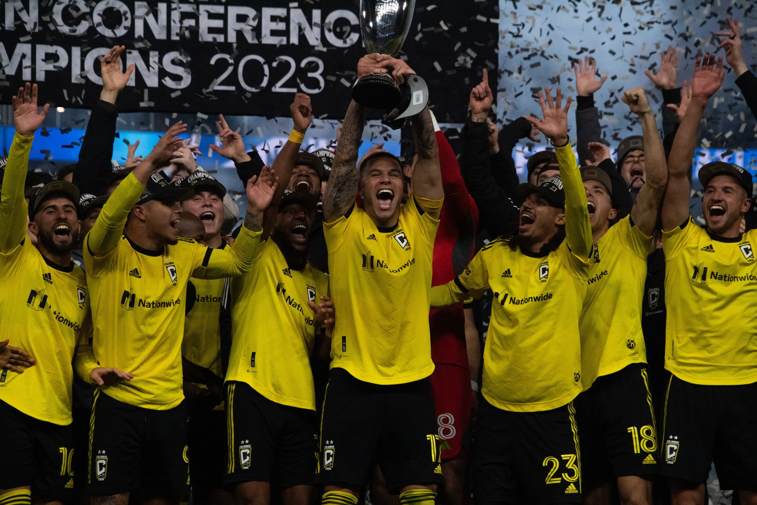 A group of soccer players in yellow jerseys celebrating a victory on a stage, some with raised hands, with a player in red holding a trophy above his head surrounded by confetti.