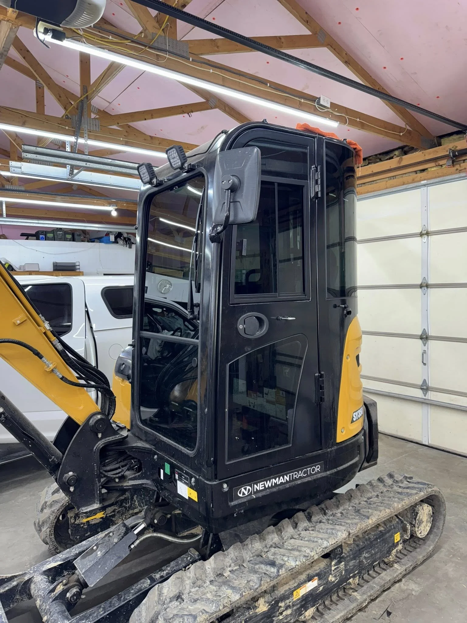 A small construction excavator inside a garage, with a black cab, yellow arm, and tracks, labeled 'Newman Tractors'.