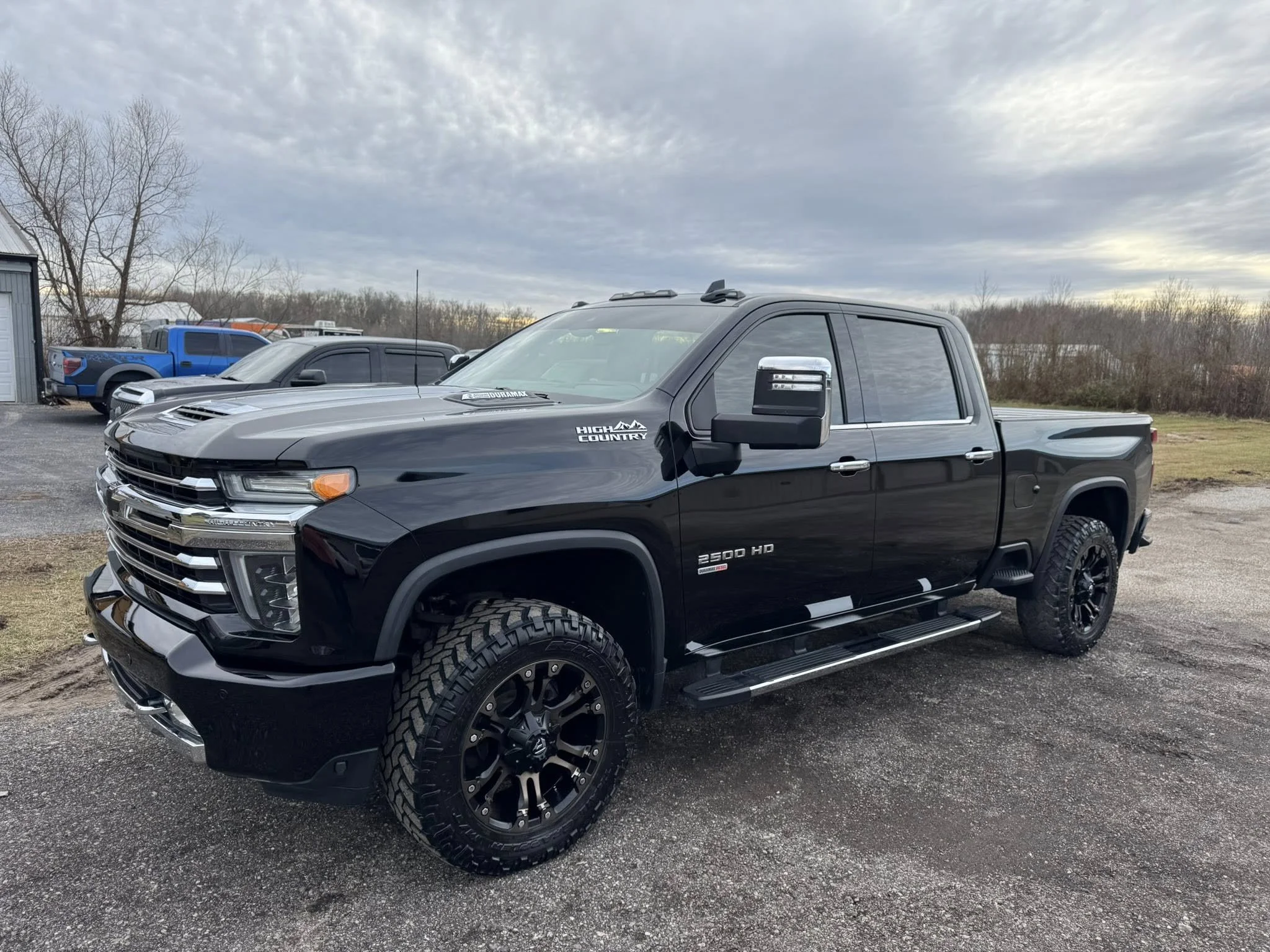 Black Chevrolet Silverado 2500 HD pickup truck with off-road tires and custom mirrors, parked outdoors on a gravel lot