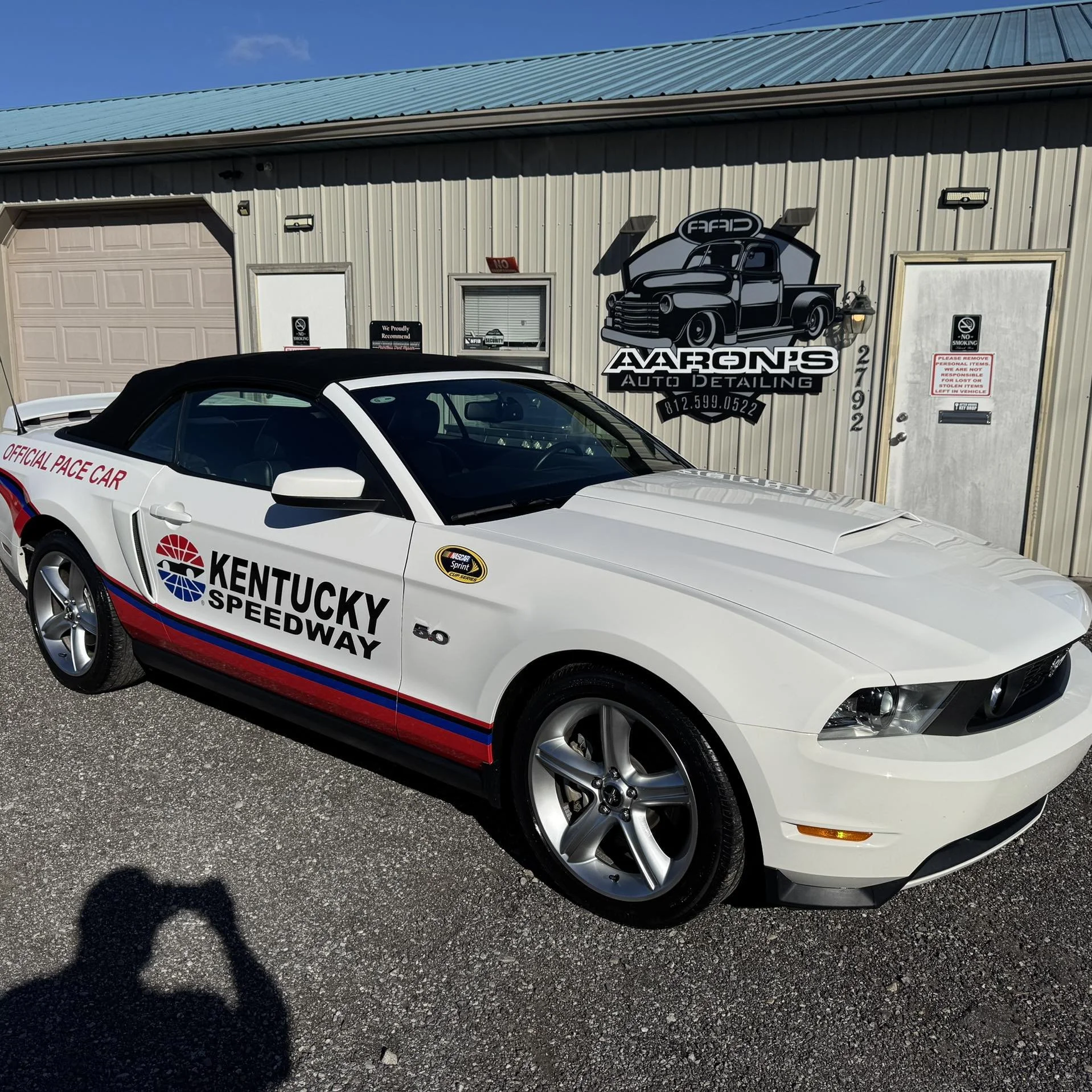 A white Ford Mustang race car with 'Kentucky Speedway' and 'Official Pace Car' decals, parked outside a detailing shop called Aaron's Auto Detailing.