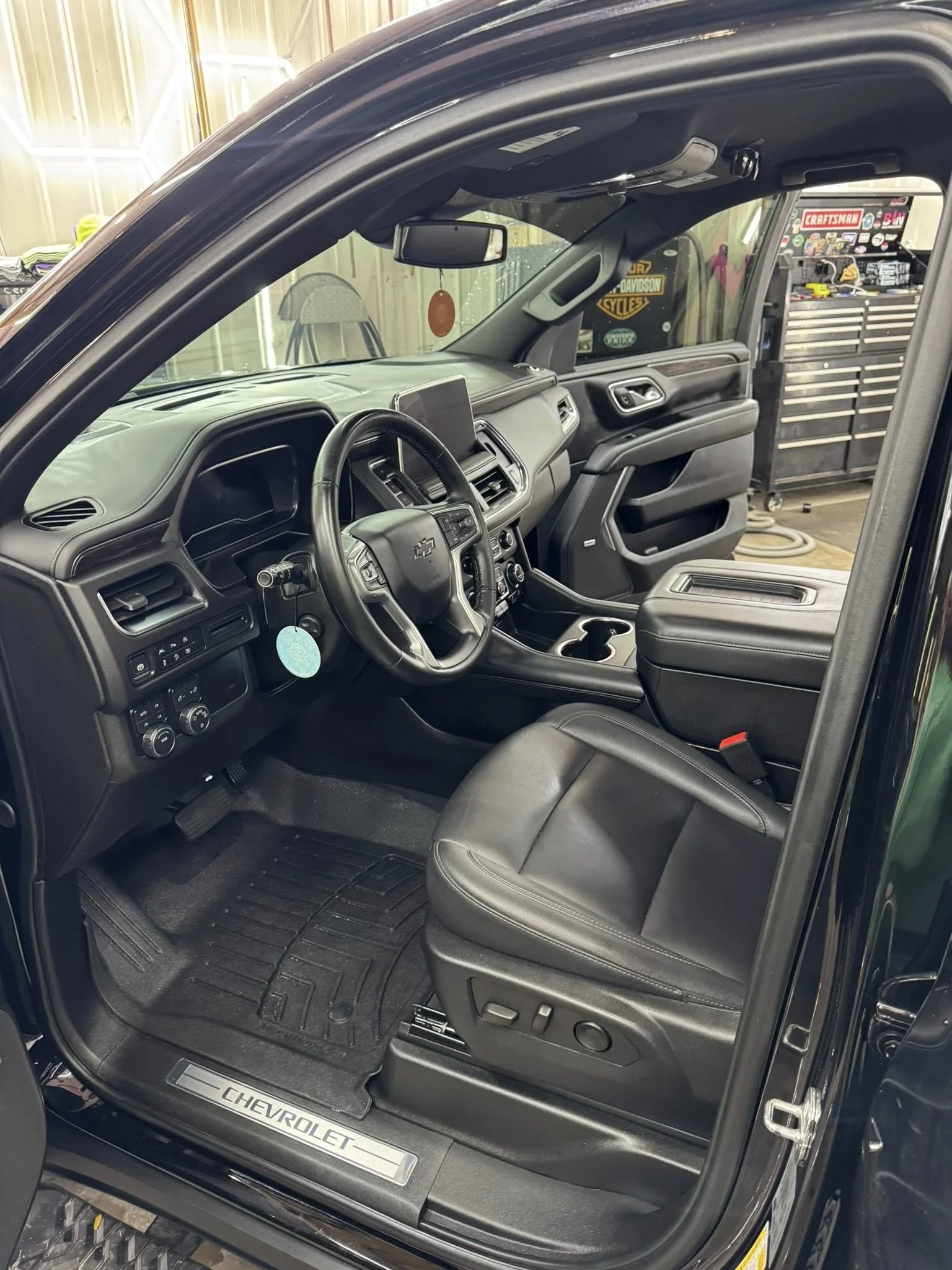 The interior of a black Chevrolet truck, showing the dashboard, steering wheel, front seat, and center console in a garage with tools and equipment in the background.