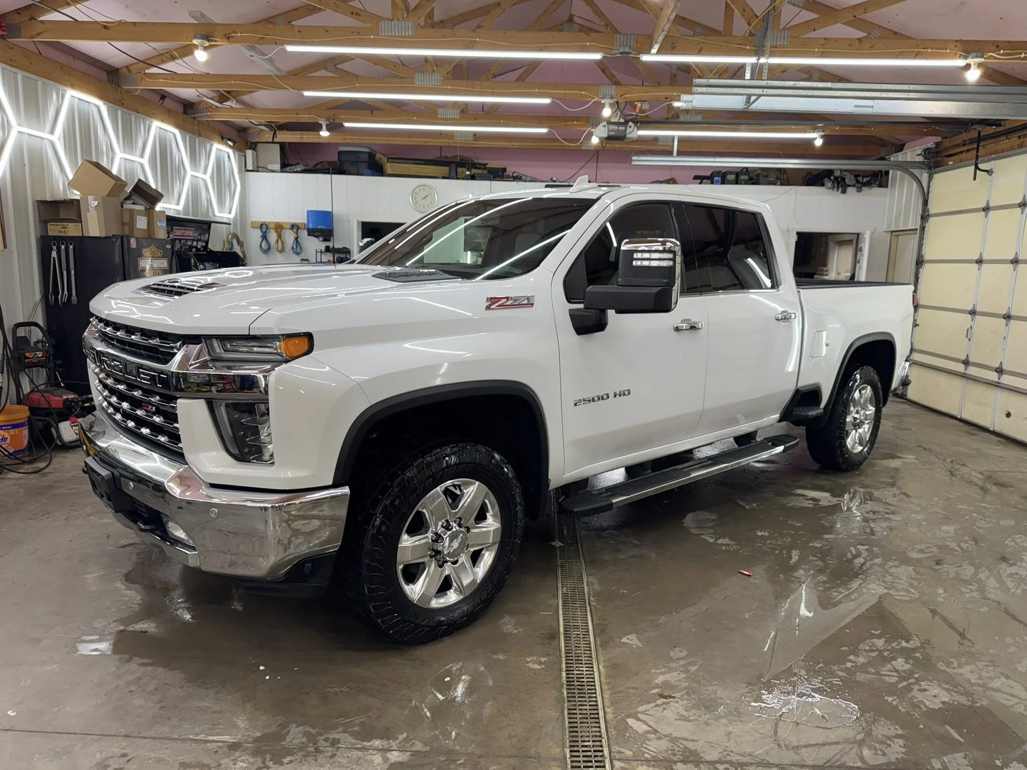 A white Chevrolet Silverado ZR2 pickup truck parked inside a garage with wet concrete floor, tools, and shelves on the walls.