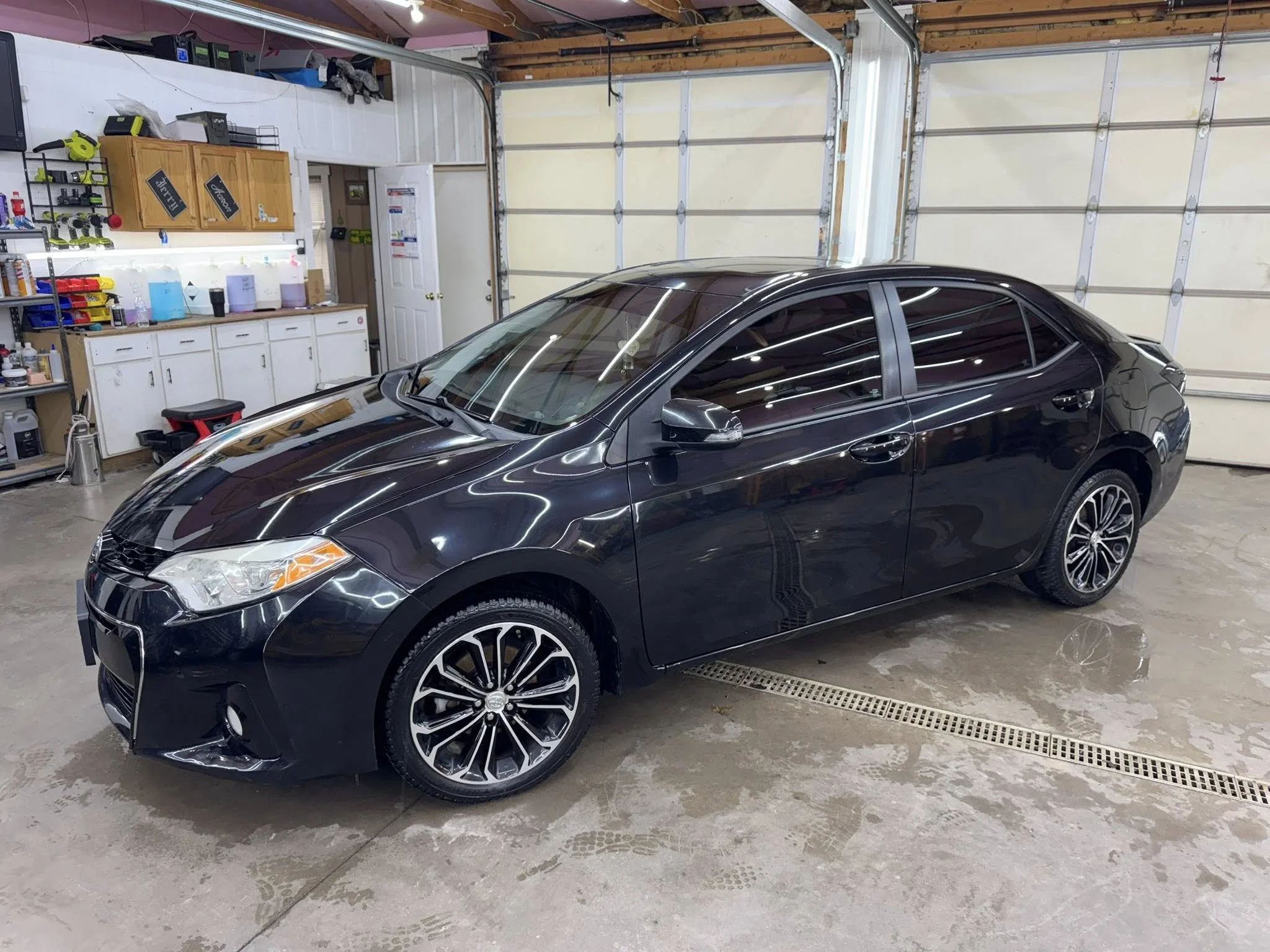 A black sedan parked inside a garage with white cabinets and shelves in the background, and fluorescent lighting reflecting off the car's surface.