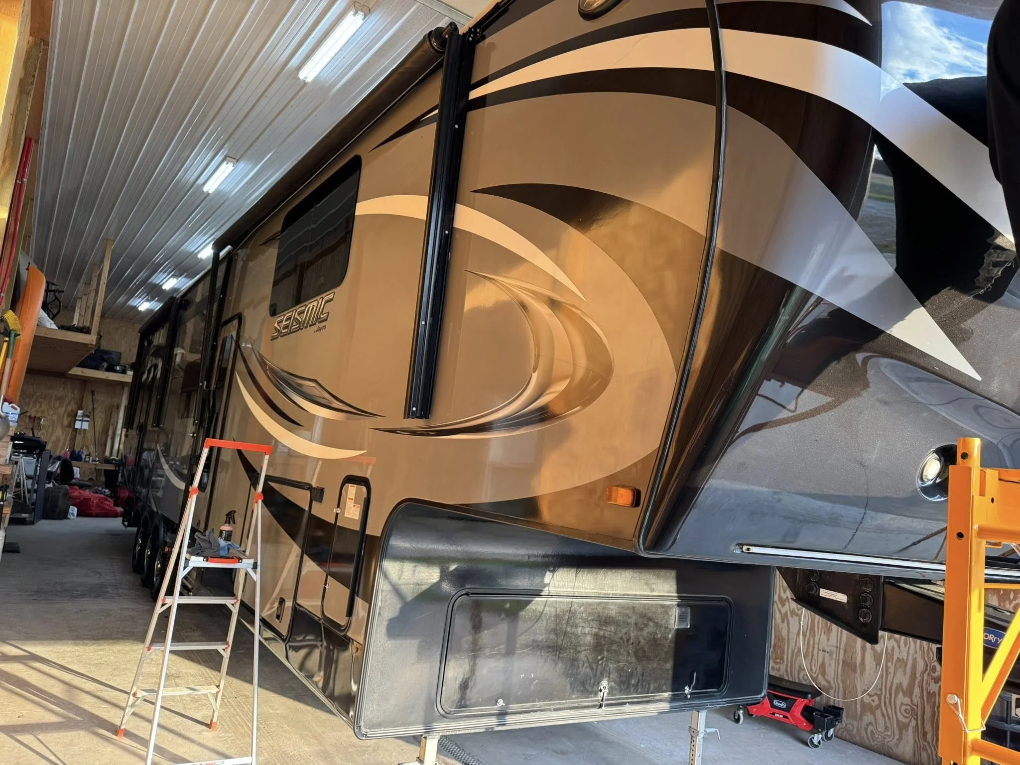 A large, shiny, multicolored Seismic travel trailer inside a wooden garage or workshop with tools, ladder, and equipment around it.