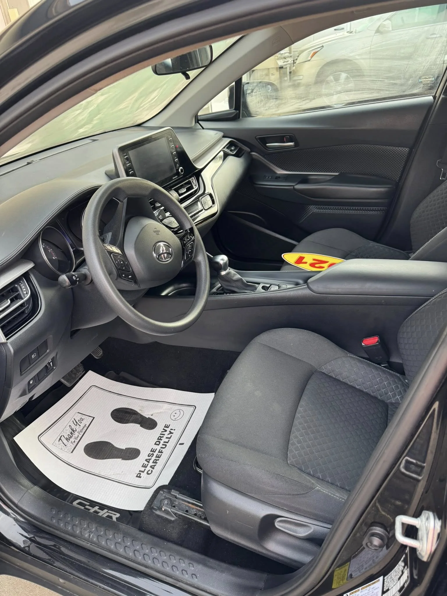 Inside of a modern car showing the driver's seat, steering wheel, dashboard, center console, and passenger seat. A paper floor mat with footprints and the message 'Please Drive Carefully!' is on the driver's side floor.