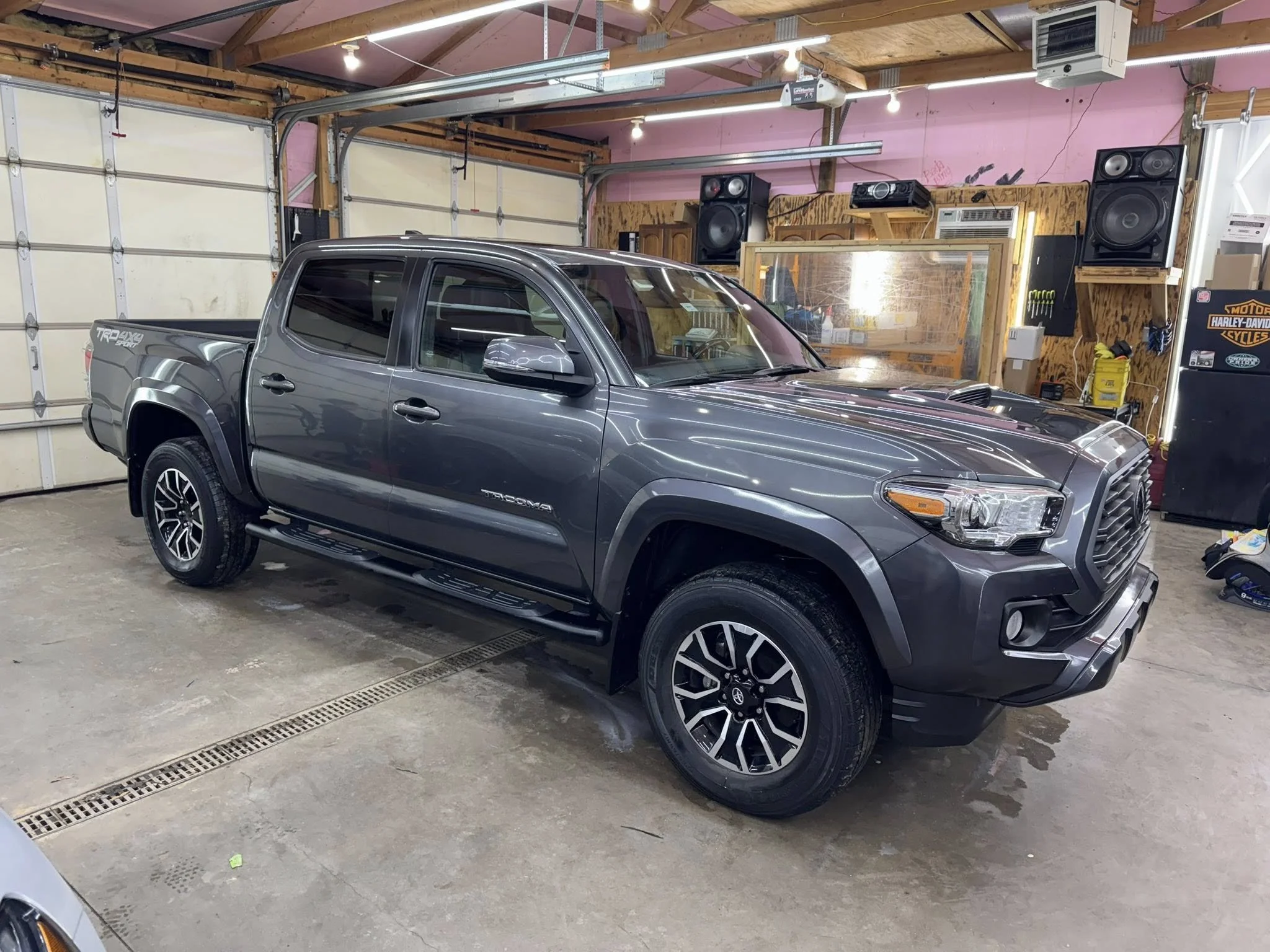 A grey Toyota Tacoma pickup truck parked inside a garage with a concrete floor, pink walls, and various tools and speakers on the walls.