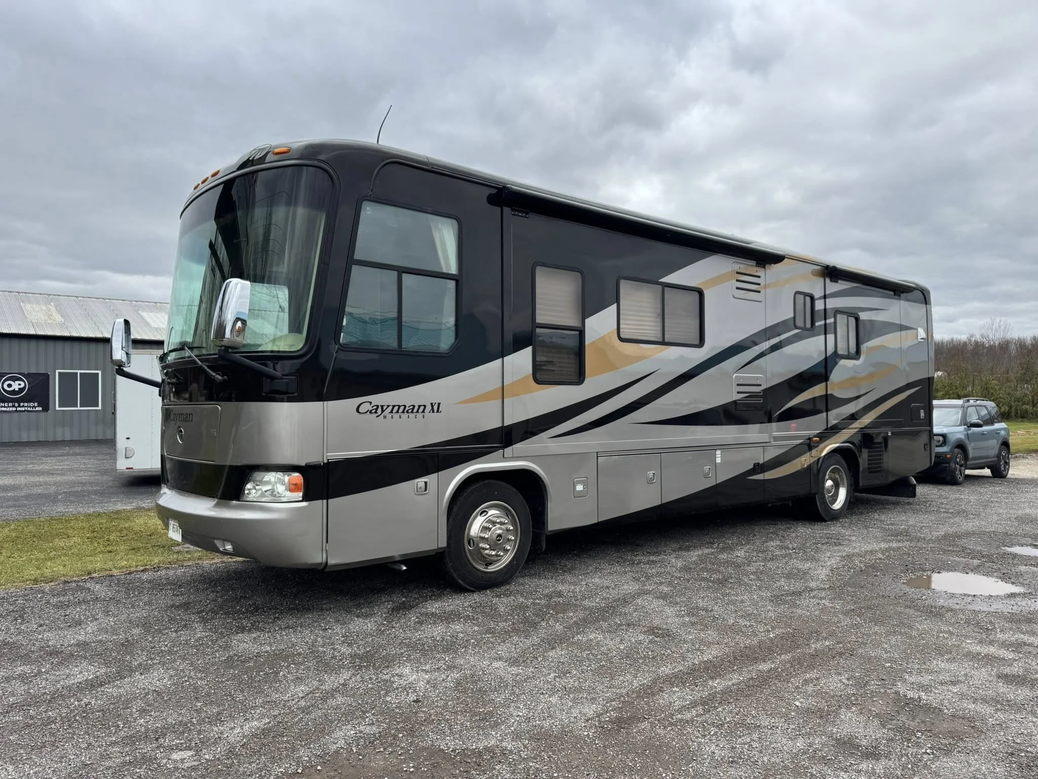 A large black and gray recreational vehicle (RV) with gold and silver accents parked on a gravel lot, with a cloudy sky overhead.