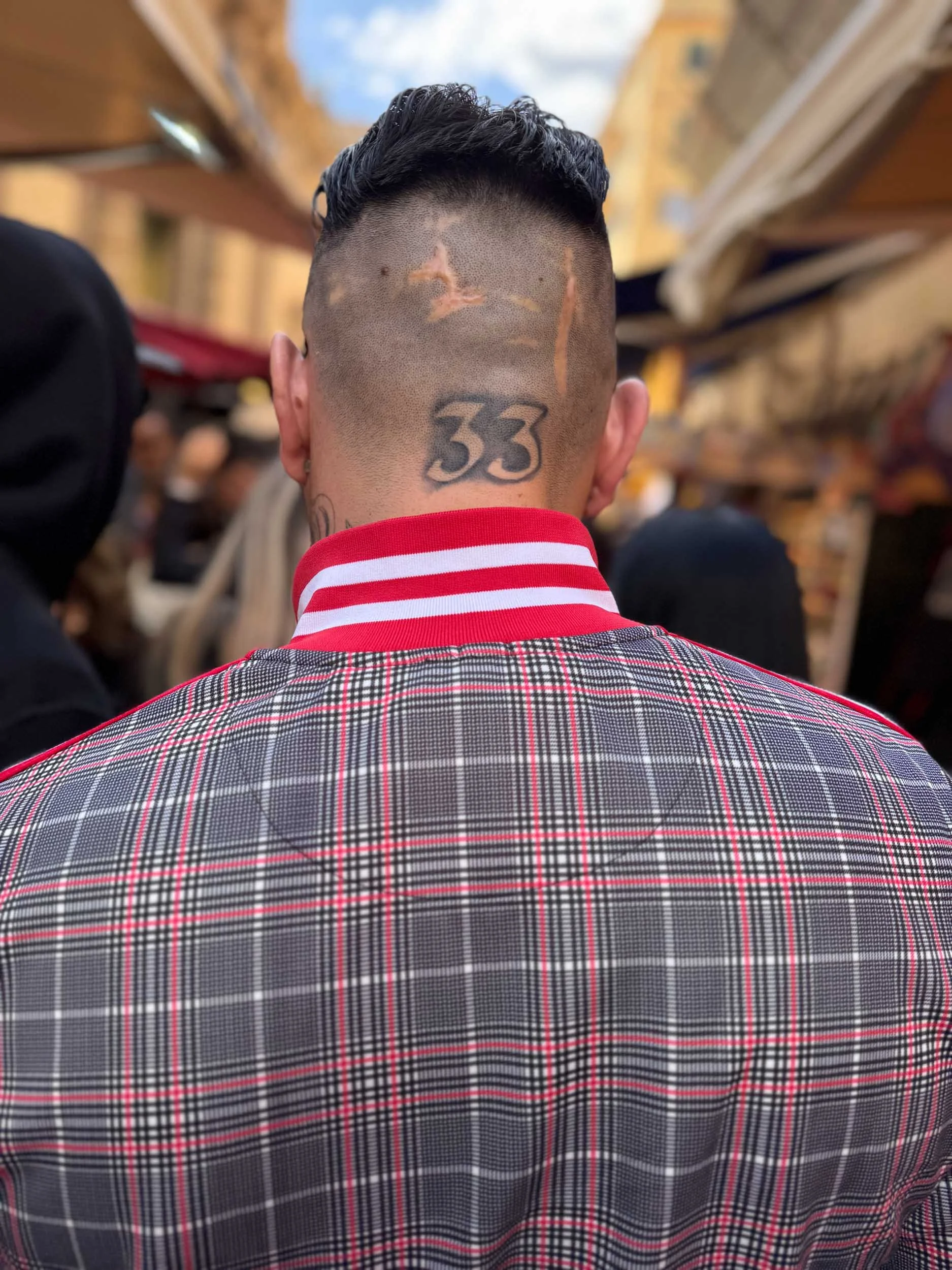 Color photo taken by Donna Lancia in Palermo of a dorsal view of a man with an interesting haircut, scars and tattoo walking in a busy market.