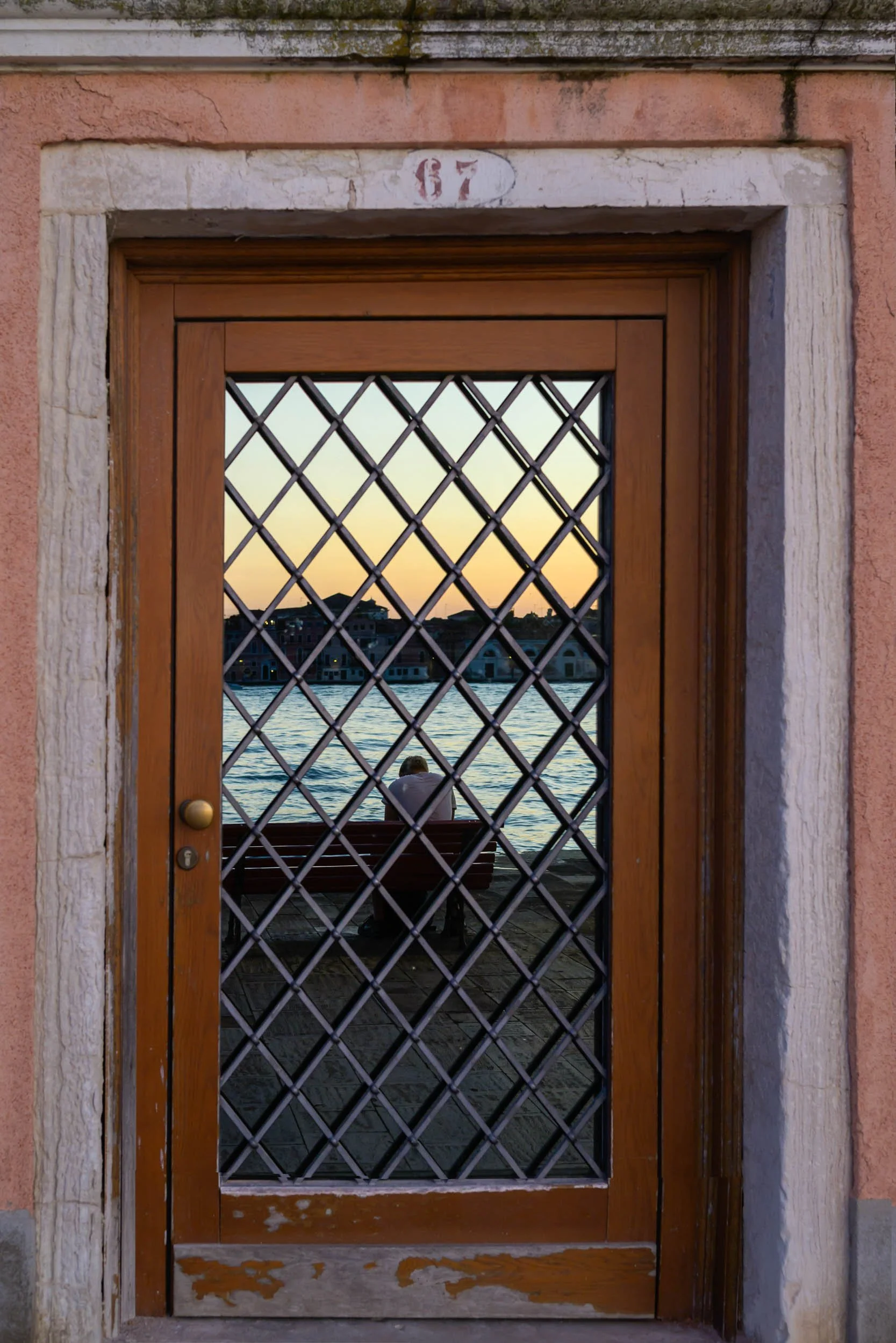 Colour photo taken by Donna Lancia in Venice of a reflection in a graphical doorway with a young man sitting on a bench at sunset.