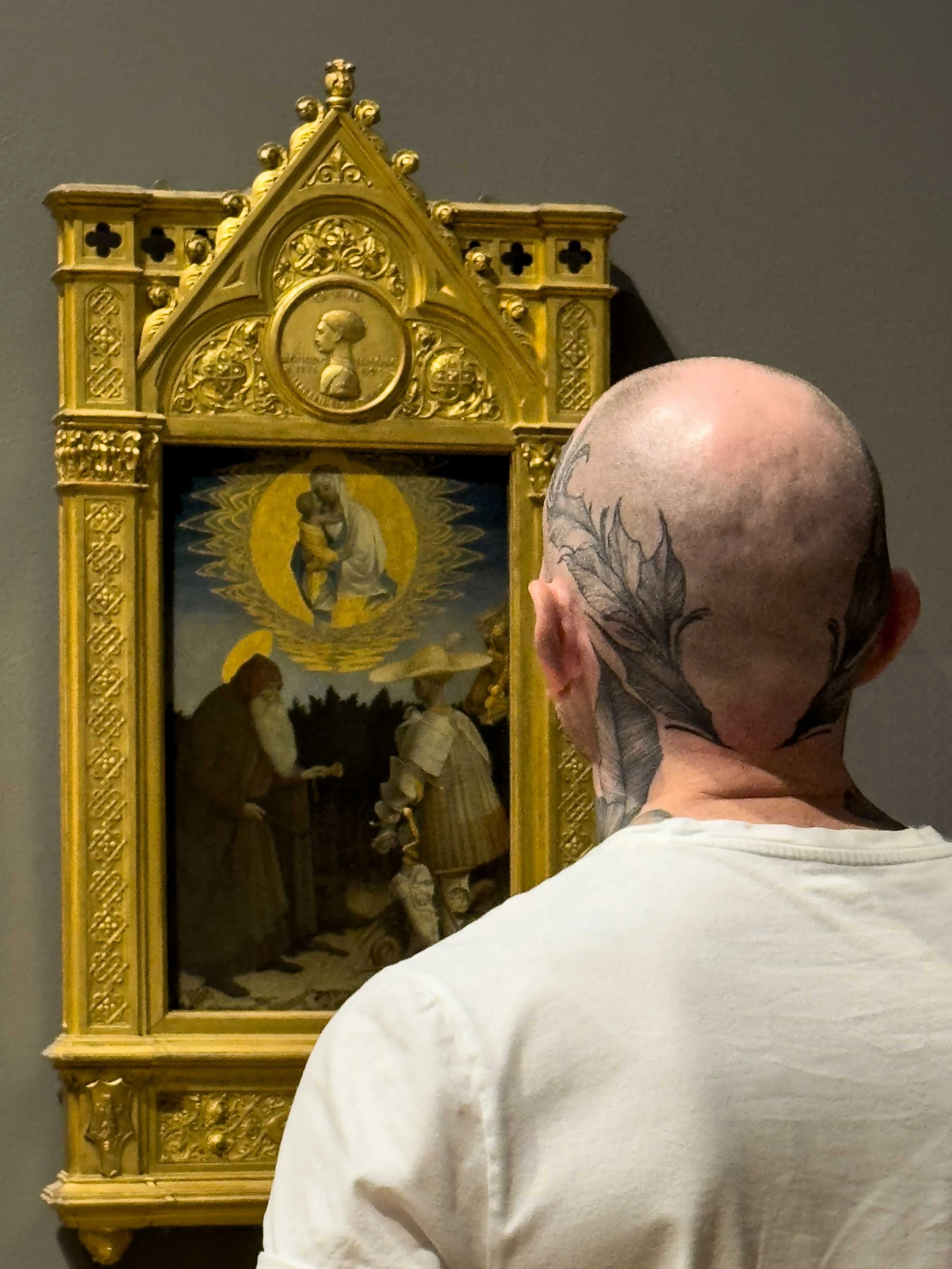 Color photo taken in London with an iPhone of a dorsal view of man with an interesting tattoo looking at religious medieval painting in the National Gallery.