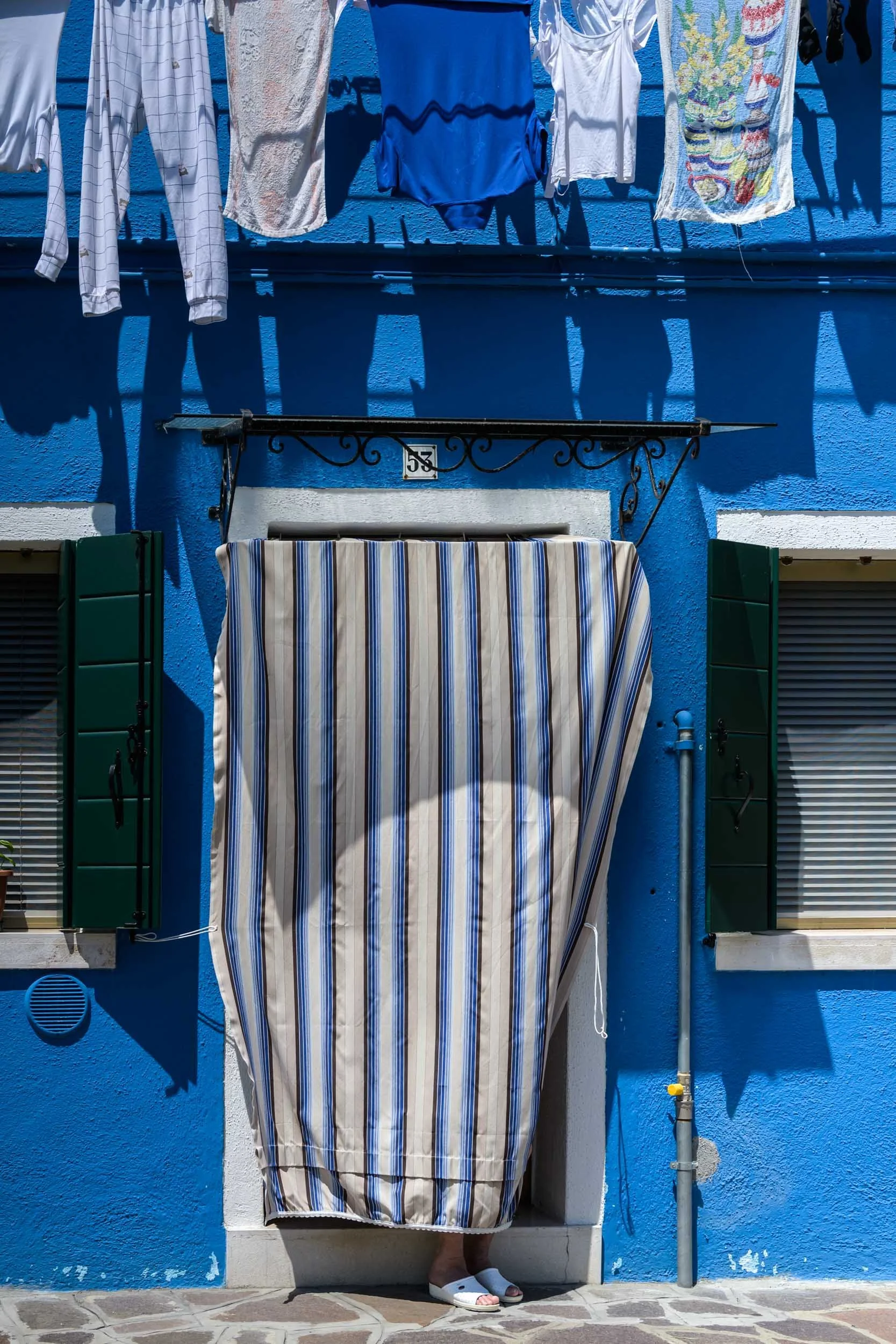 Colour photo taken by Donna Lancia in Burano, Italy of blue house with matching blue laundry and a lone woman sitting on the stoop where only her feet are visible.