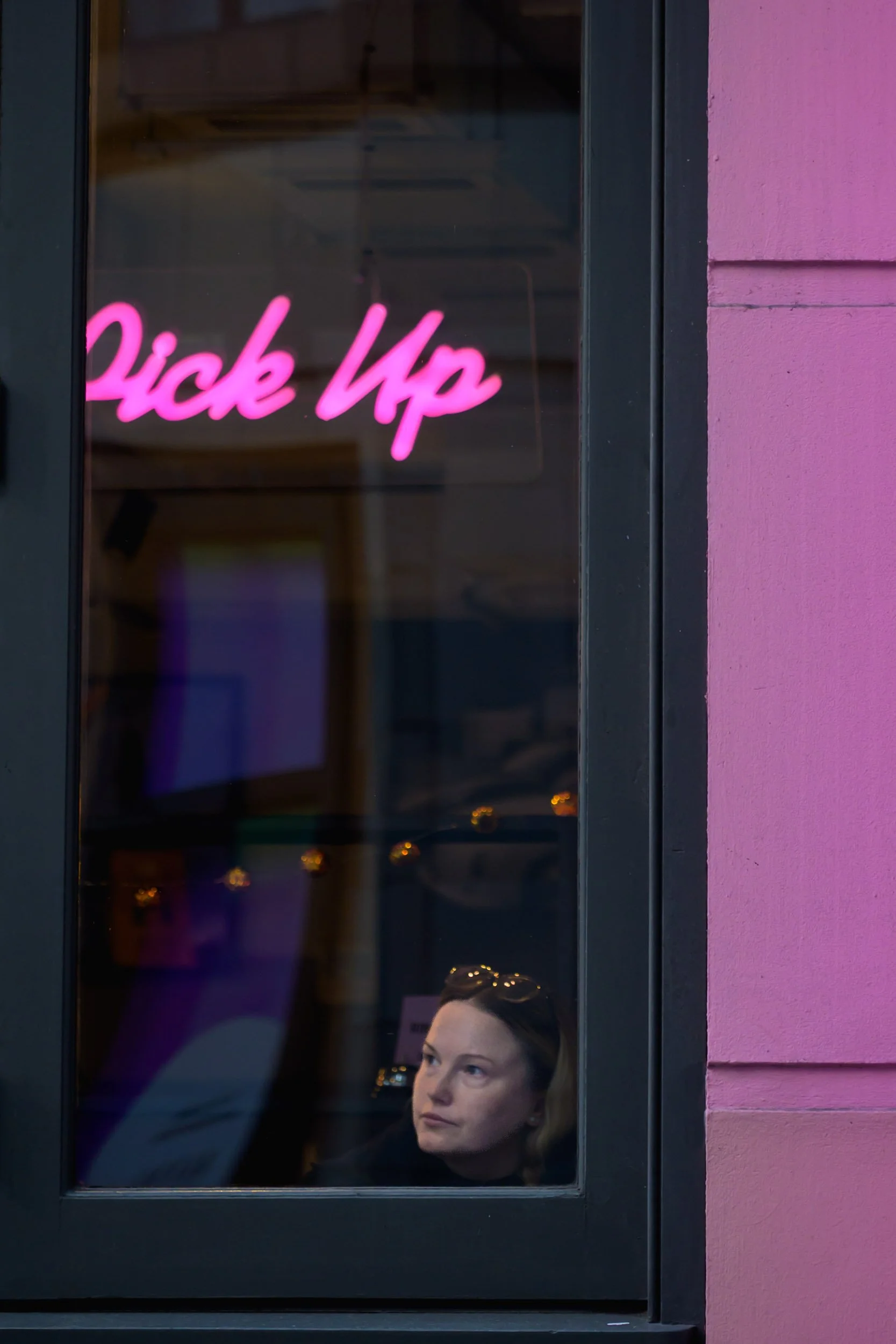 Colour photo taken by Donna Lancia in Soho, London of a girl looking forlornly out of a shop window with the word Pick Up in pink matching the wall outside..  