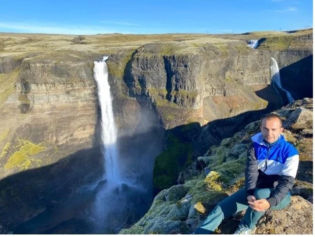 Persona sentada en rocas frente a una cascada en un cañón, con un cielo despejado.