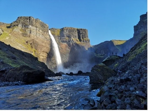Cascada en un cañón con rocas y agua corriente en primer plano, y paredes rocosas altas con vegetación escasa bajo un cielo despejado.