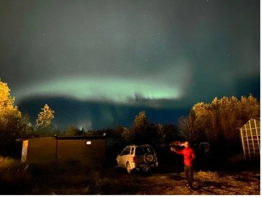Aurora boreal en la noche sobre un paisaje con árboles, autos y una persona tomando fotos.