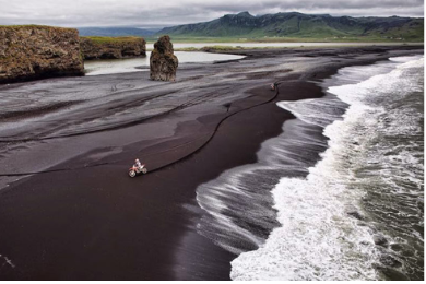 Playa de arena negra con una persona en un cuatriciclo cerca del mar y formaciones rocosas en el fondo.