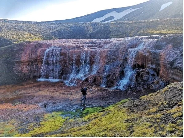 Una persona explorando cerca de una cascada en un paisaje volcánico con nieve en la cima.