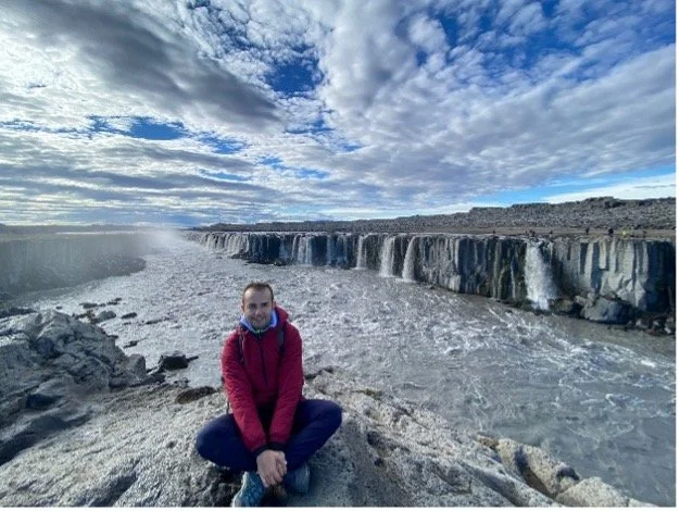 Persona sentada en la orilla de un río con cascadas y cielo nublado en el fondo.
