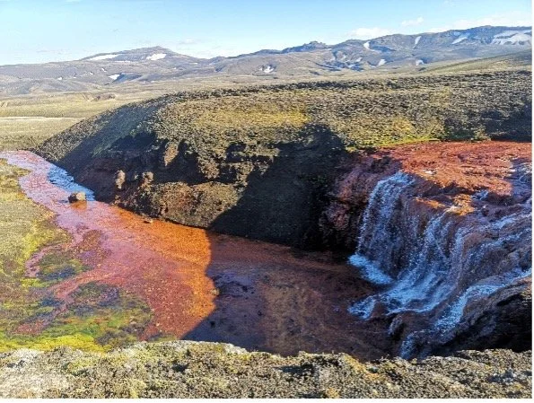 Cascada en un cañón con río colorido y paisaje montañoso al fondo.