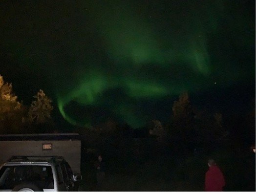 Auroras boreales verdes en el cielo nocturno sobre un área con árboles, una coche y una persona observándolas.
