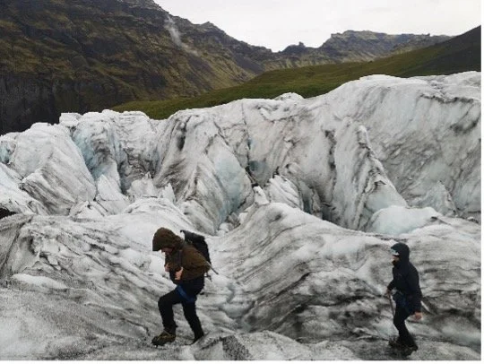 Dos personas caminando sobre un glaciar con montañas y praderas verdes en el fondo.