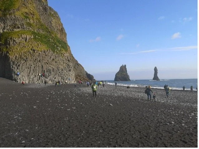 Playa de piedras negras con formaciones rocosas en el mar y personas caminando, junto a un acantilado con vegetación.