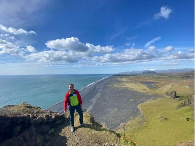 Persona de pie en un acantilado con vista a una playa y el océano, paisaje con cielo azul y nubes.