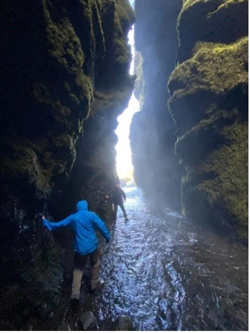 Personas explorando una cueva estrecha con agua en el suelo en un entorno natural rocoso y con luz brillante al fondo.