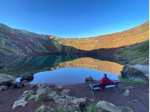 Persona sentada en un banco junto a un lago en un paisaje de montaña, con cielo azul despejado.