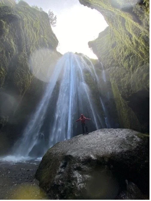Persona de pie sobre una roca grande en un cañón con una cascada que cae desde una cueva en la parte superior, rodeada de paredes cubiertas de musgo y vegetación.