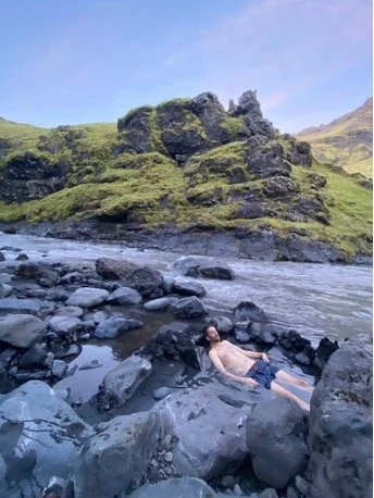 persona descansando sobre rocas cerca de un río en un paisaje montañoso con formaciones rocosas y vegetación verde