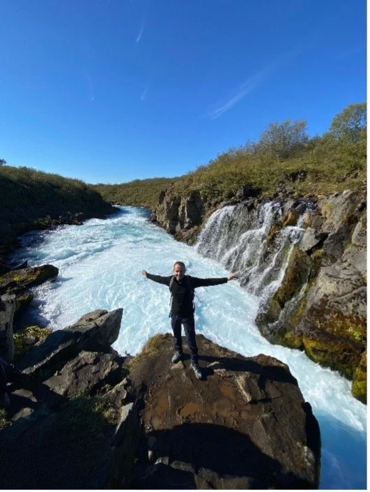 Persona de espaldas, con brazos extendidos, de pie sobre una roca, cerca de un río con cascadas, en un paisaje de naturaleza y cielo despejado.